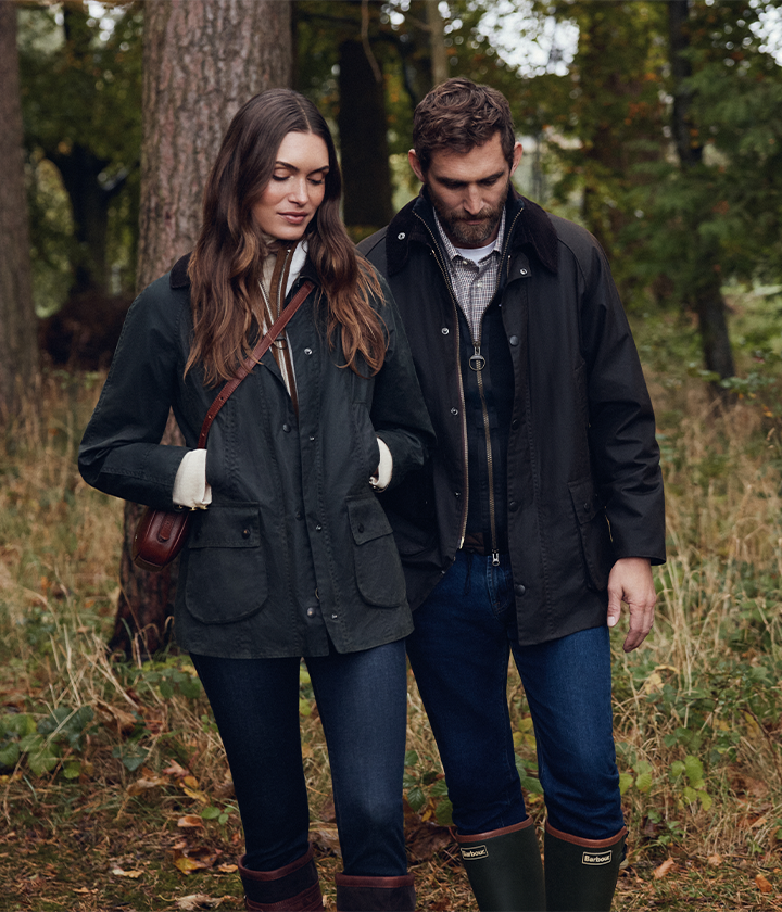 A couple wearing dark waxed jackets and jeans walks through a wooded area with autumn foliage.